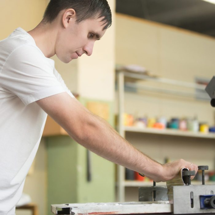 Young attractive worker man working using printmaking tools, silk screen printing on clothing fabric technique